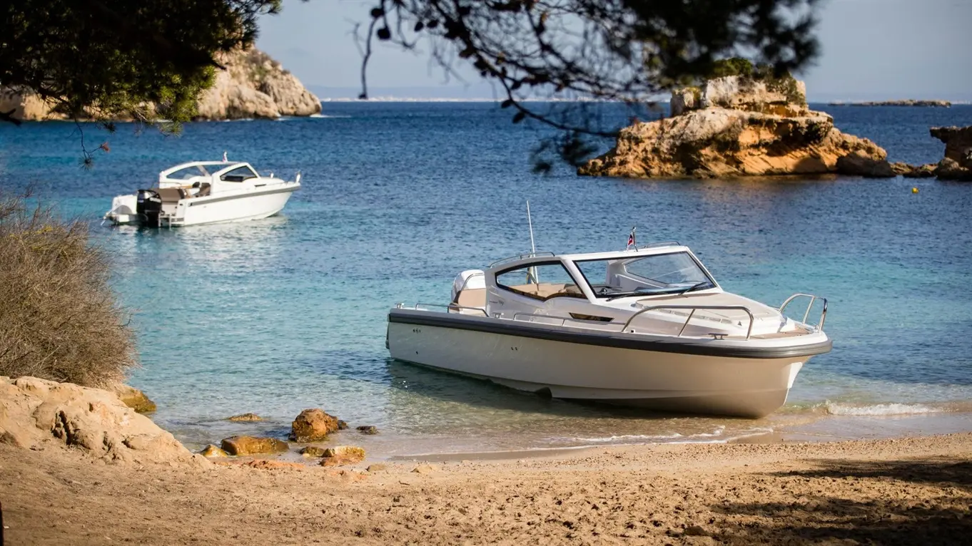 Two boats anchored in clear blue water by a sandy beach, rocky shoreline in the background, surrounded by trees.
